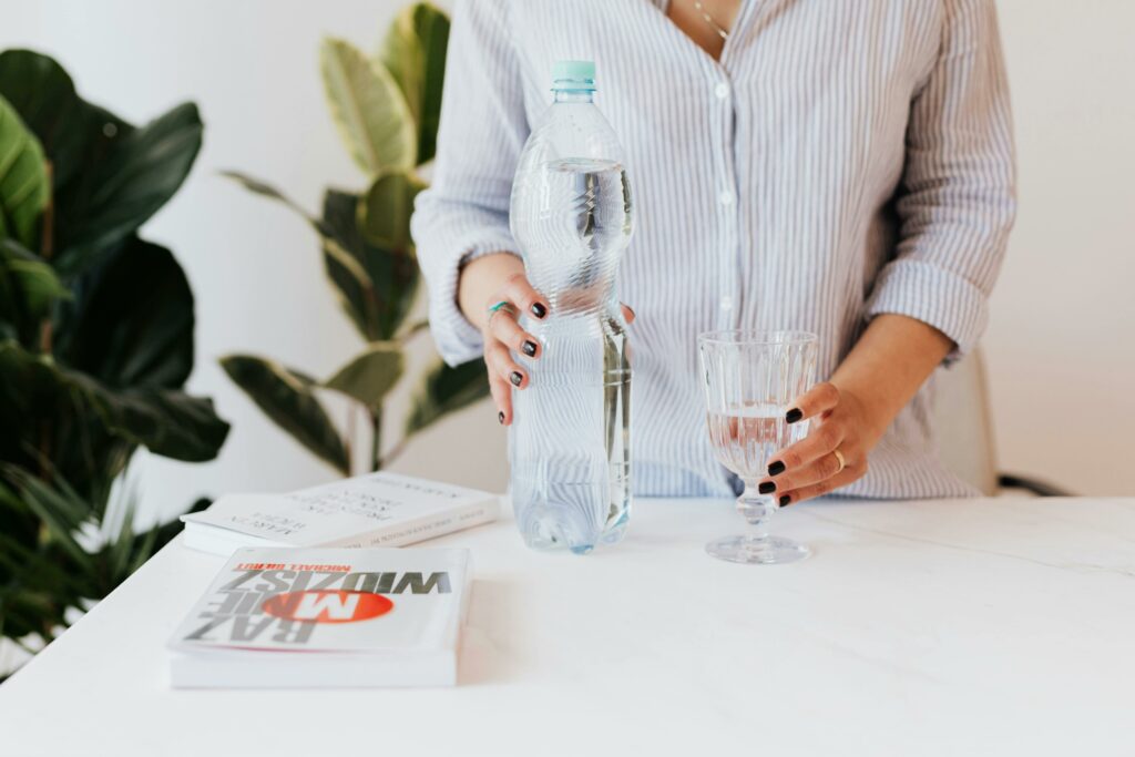 Woman holding bottle and glass of water on desk in home office with a book lying near